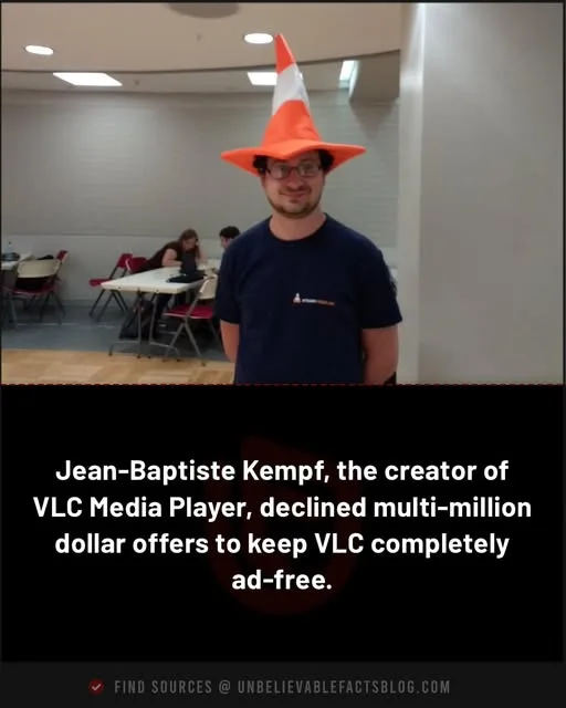 A man wearing an orange and white traffic cone hat smiles at the camera while standing indoors. In the background, other…