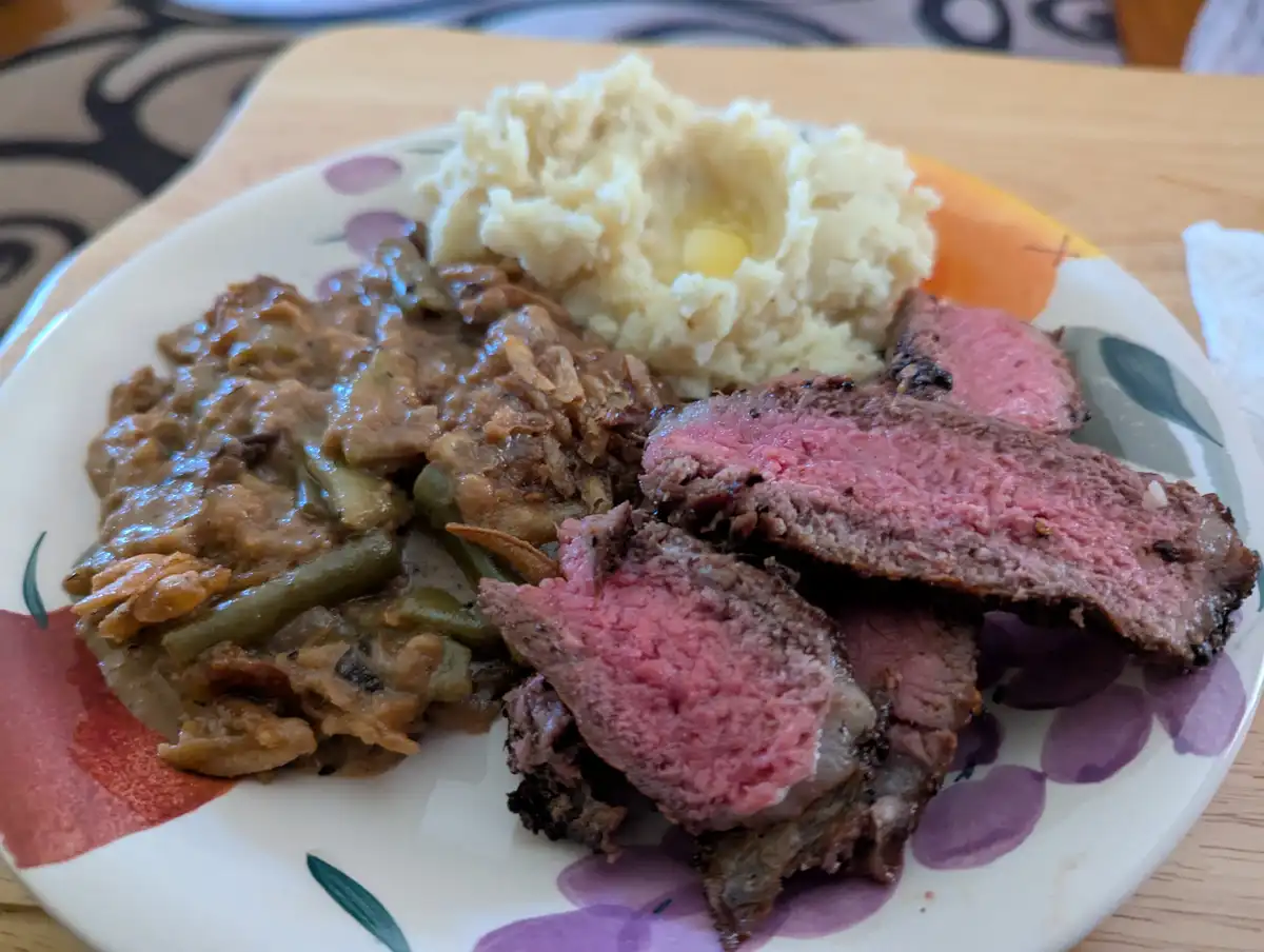 A plate of food featuring sliced medium-rare steak, mashed potatoes with a pat of butter, and green beans in a brown sauce…
