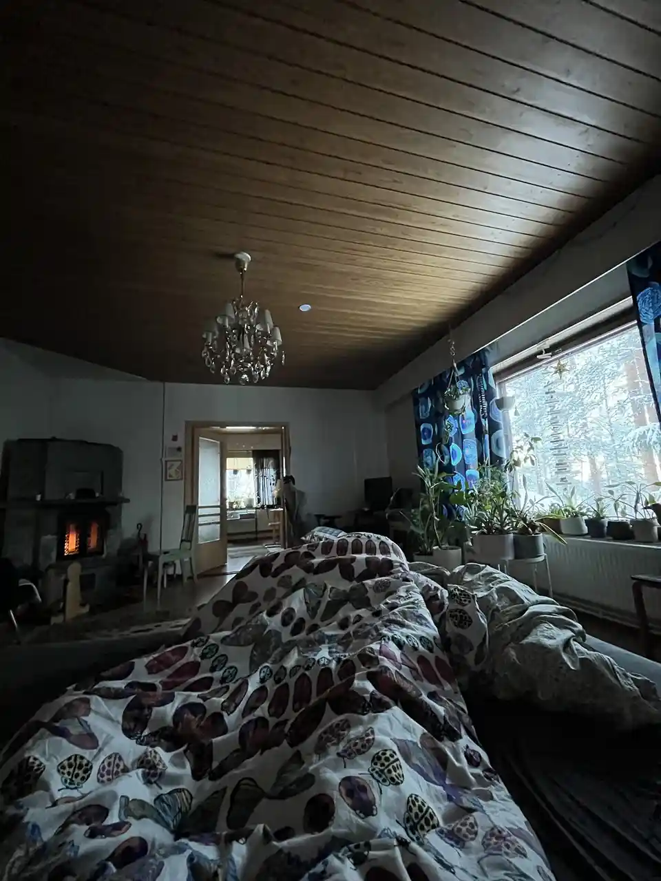 A cozy, dimly lit bedroom with a rumpled bed in the foreground, a lit fireplace in the background, and a window showing a…
