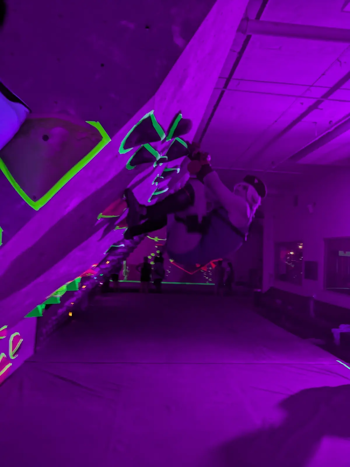 A person is hanging upside down from a climbing wall in a dark room bathed in purple light, with glowing green handholds…