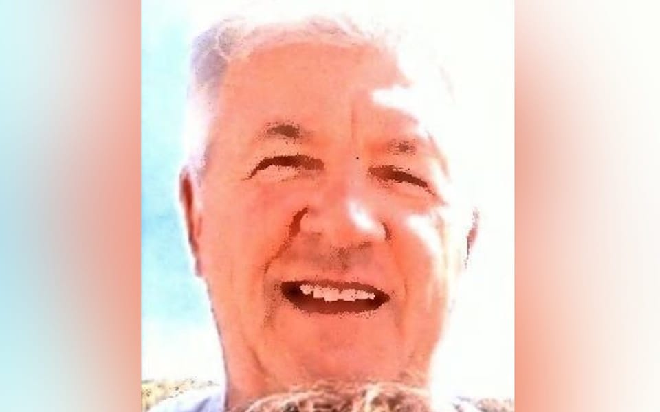 A close-up photo of an older man with white hair, smiling broadly with his eyes crinkled, taken outdoors under bright…