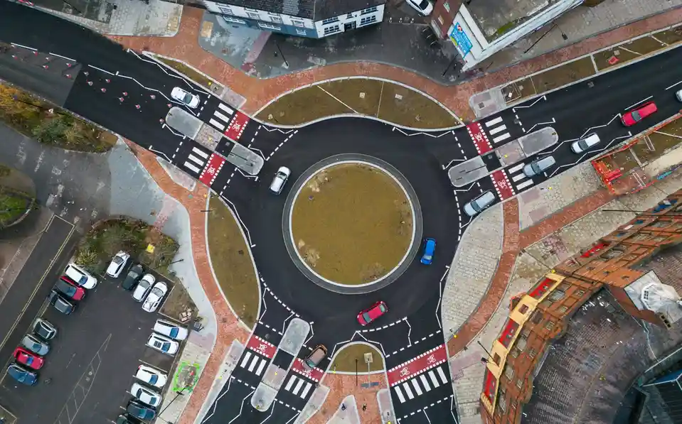 An aerial view of a modern roundabout with cars navigating its circular path, surrounded by roads, sidewalks, and buildings…