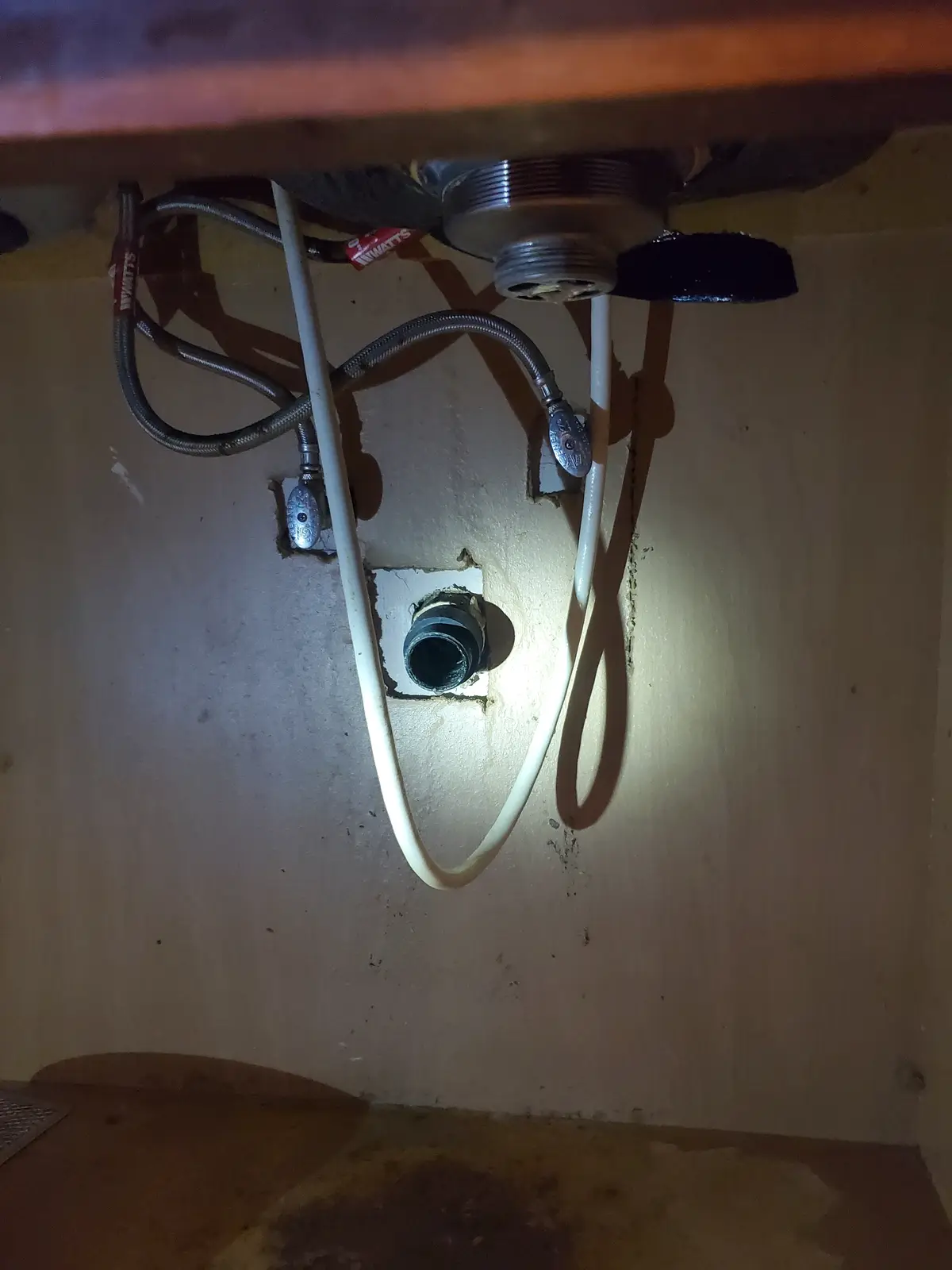 A dimly lit photo shows plumbing under a sink, with white pipes and metal braided hoses connected to valves. A white pipe…