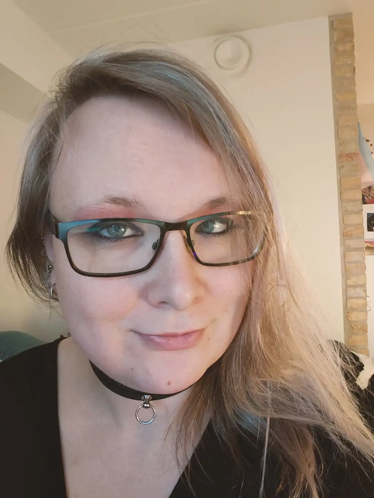 A selfie of a woman with glasses, light brown hair, and a black choker necklace, smiling slightly while looking at the…