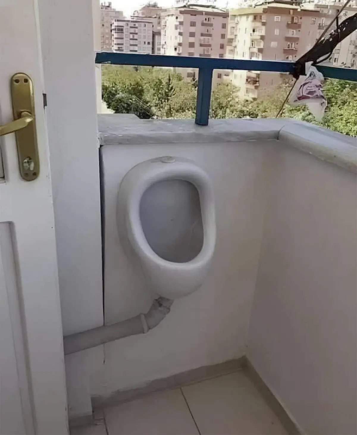 A white ceramic urinal is mounted on the interior wall of a small balcony, with a pipe extending downward. Outside, a blue…