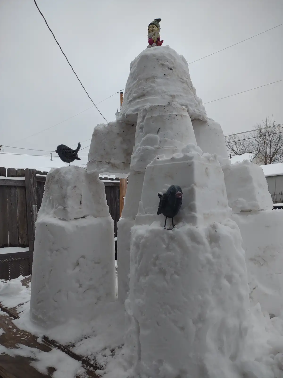 A large, multi-tiered snow fort built outdoors on a wooden deck, with a garden gnome figurine perched on top and two…