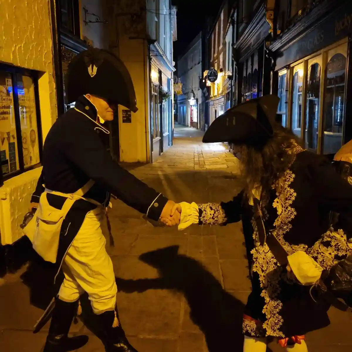 Two men in historical costumes shake hands on a narrow, lit street at night. One wears a navy officer's uniform with a…