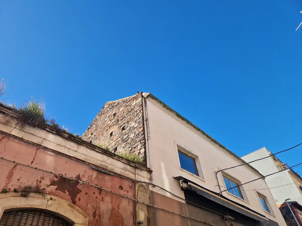 A low-angle photo shows the corner of weathered buildings under a clear blue sky, with a stone tower section and faded pink…