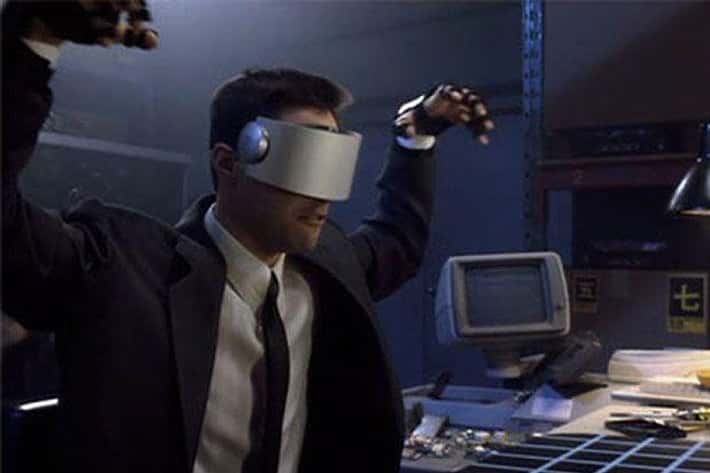 A man in a suit and tie wears a bulky, silver virtual reality headset that covers his eyes, with his arms raised in a…