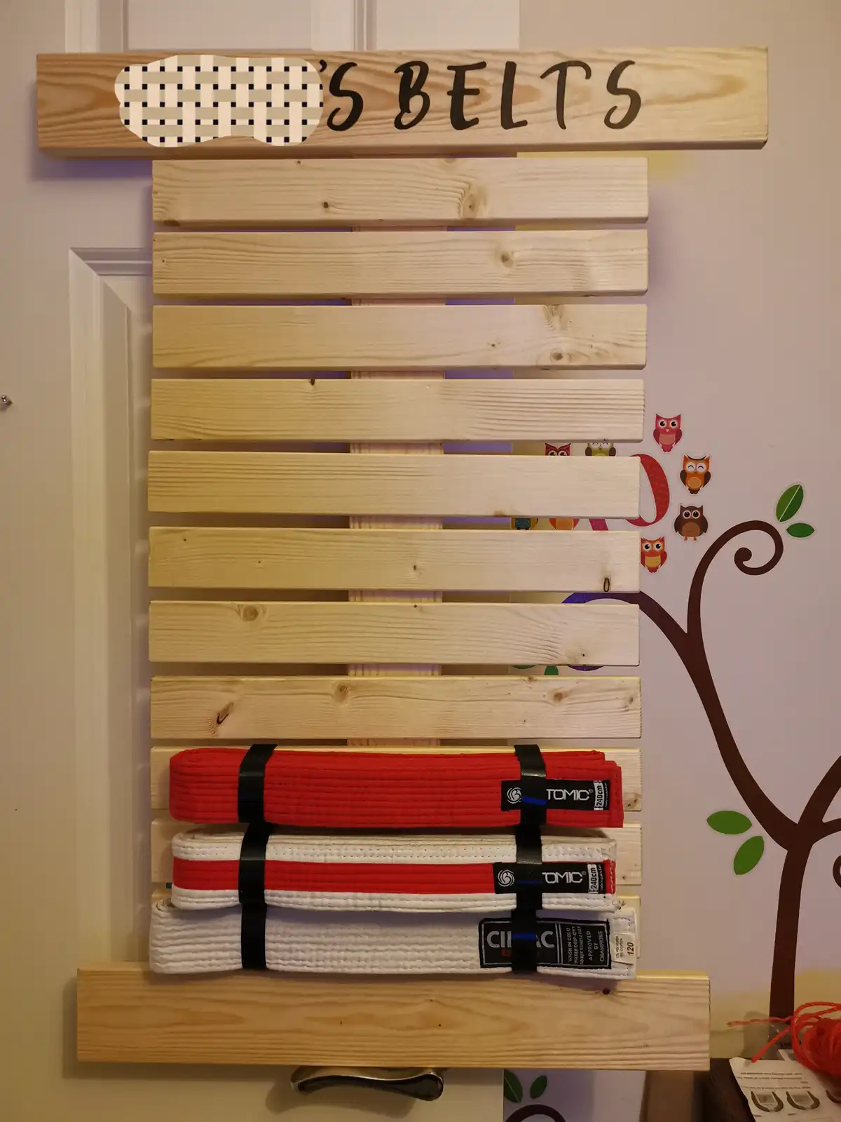 A wooden wall-mounted rack labeled 'S BELTS' with a placeholder for a name, displaying three martial arts belts (red…