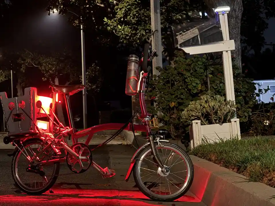 A red folding bicycle, branded 'Brompton', is parked on a sidewalk at night, illuminated by glowing red lights on its frame…