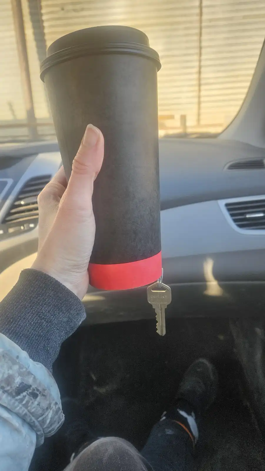 A hand holds a black disposable coffee cup with a red band near the bottom, from which a single car key dangles by a ring…