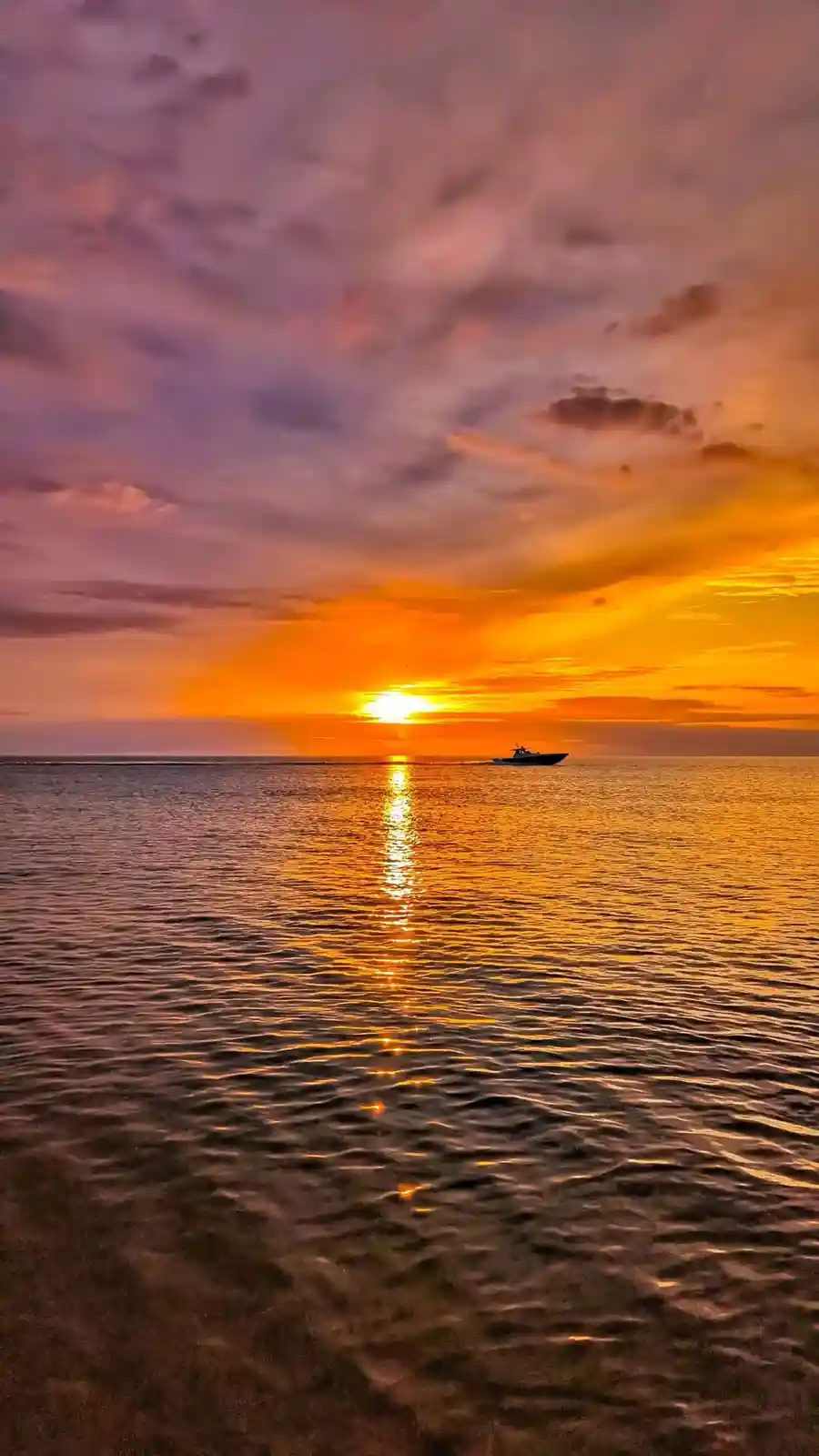 A serene sunset over the ocean with vibrant orange and purple clouds, a small boat silhouetted on the water, and the sun's…