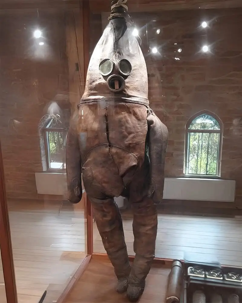 a full-body, vintage diving suit made of brown leather with a large helmet and two round glass eye pieces is hanging from a…