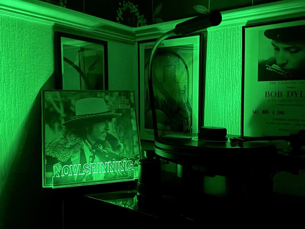 A room bathed in green light features a record player with a Bob Dylan 'Desire' album cover standing upright next to it. The…