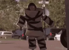 A person wearing a bulky, striped, white-and-black costume resembling a mummy or a padded suit walks toward the camera, then…