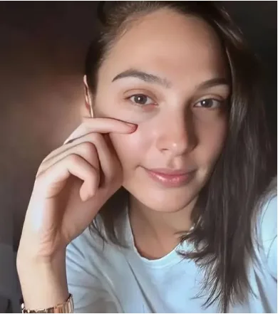 A woman with dark hair and a light blue t-shirt looks directly at the camera with a slight smile while gently touching her…