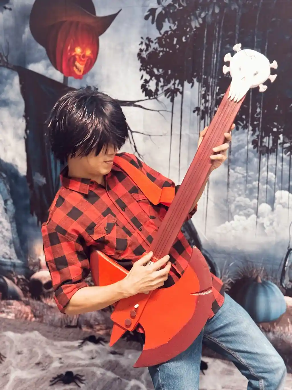 A person in a red and black plaid shirt and jeans poses holding a red, axe-shaped guitar with a skull headstock, in front of…