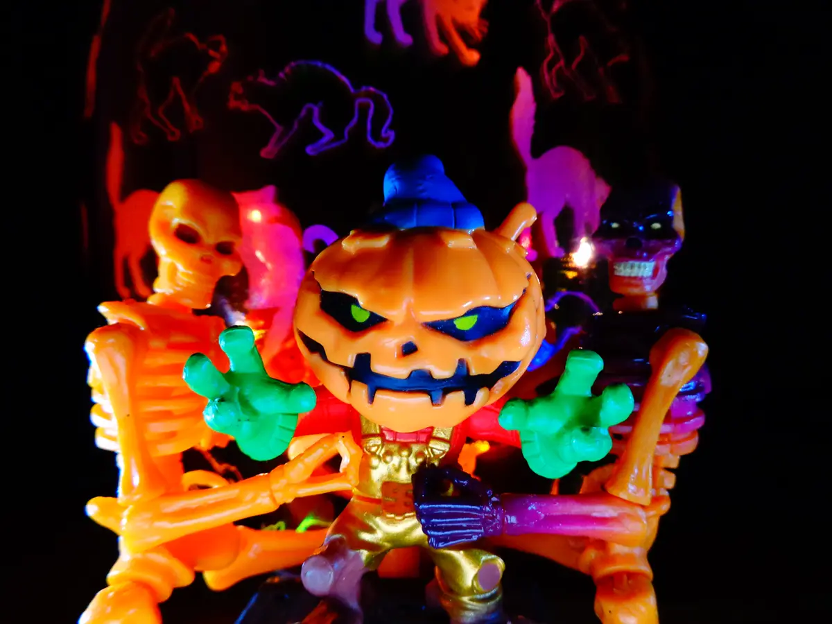 A close-up of a plastic Halloween toy featuring a central orange pumpkin-headed figure with glowing green eyes and a blue…