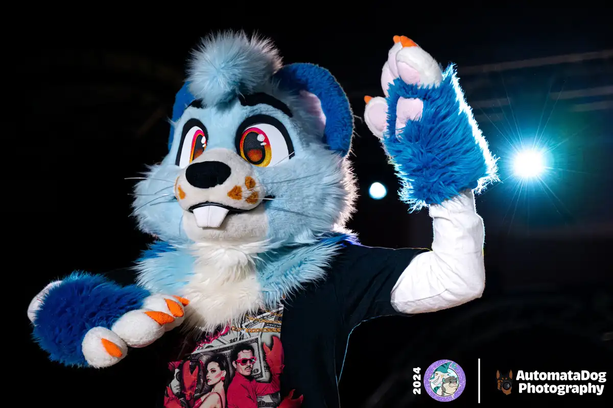 A person in a blue and white fursuit resembling a rodent or beaver, wearing a black t-shirt with a graphic print, poses with…