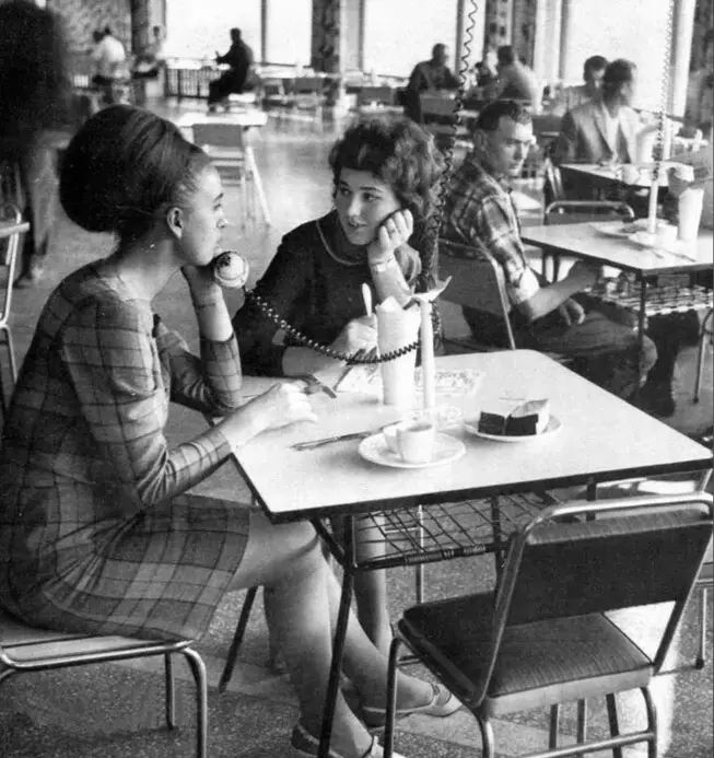 A black-and-white photo from the 1950s or 60s shows two women sitting at a diner table, each holding a rotary phone receiver…