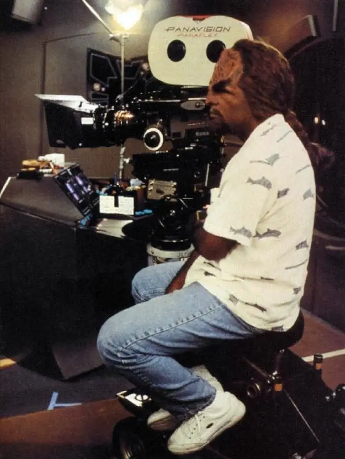 A man with long hair and a white shirt with shark prints sits on a camera dolly in front of a large Panavision Panaflex…