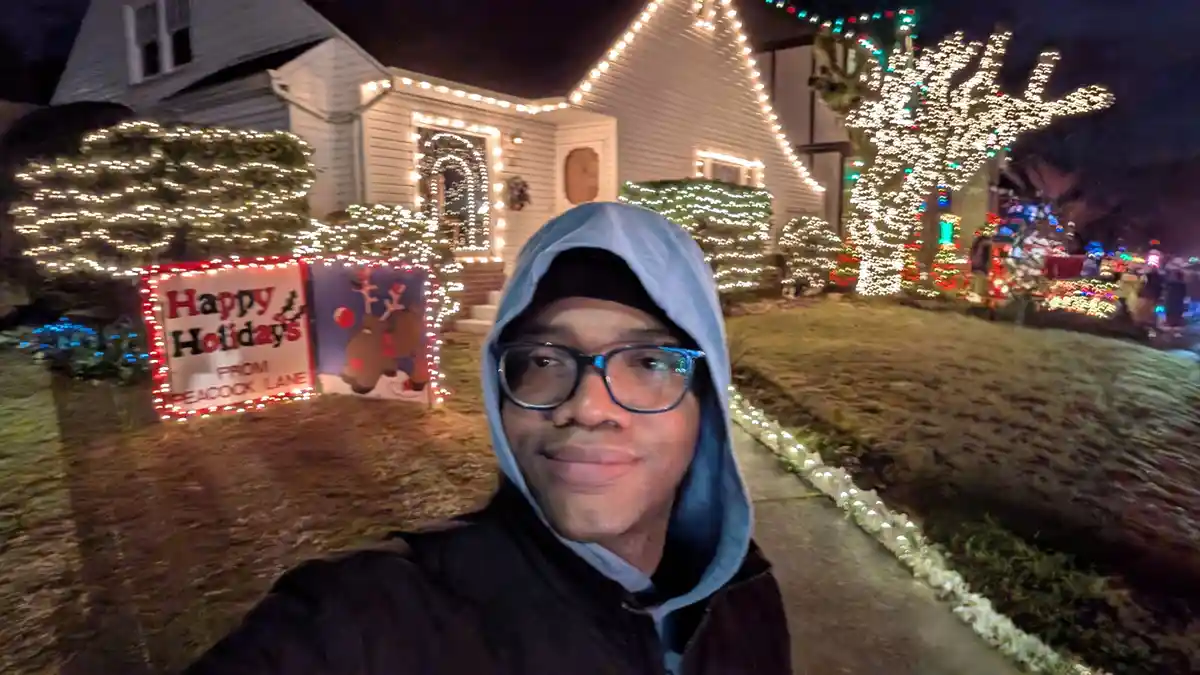 A person in a blue hoodie and glasses takes a selfie in front of a house extravagantly decorated with Christmas lights…
