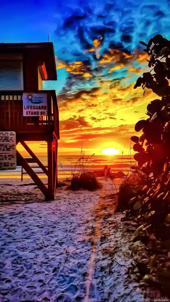 A vibrant, heavily saturated beach sunset photo featuring a wooden lifeguard stand on the left with a sign reading…