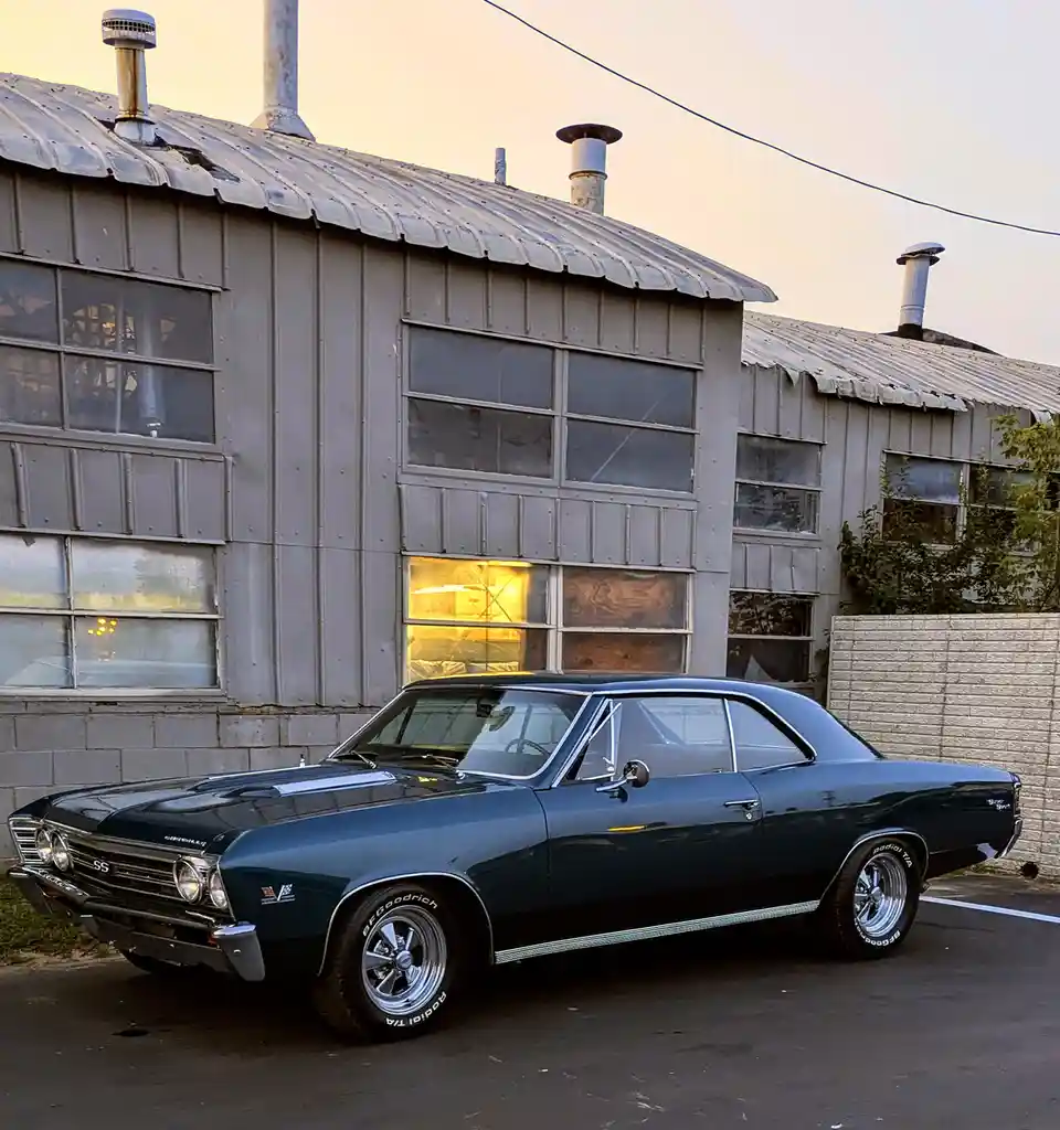 A dark blue classic 1960s Chevrolet SS muscle car with chrome trim and BF Goodrich tires is parked in front of a weathered…
