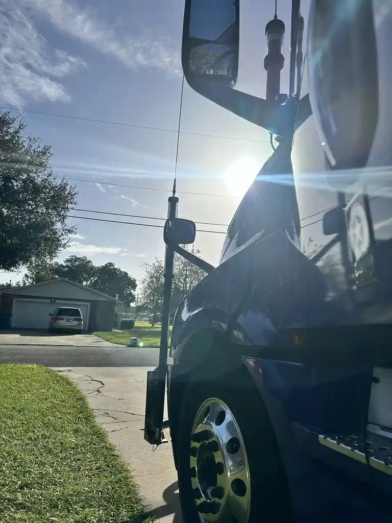 A low-angle photo shows the side of a large blue truck parked on a residential street, with the sun shining brightly behind…