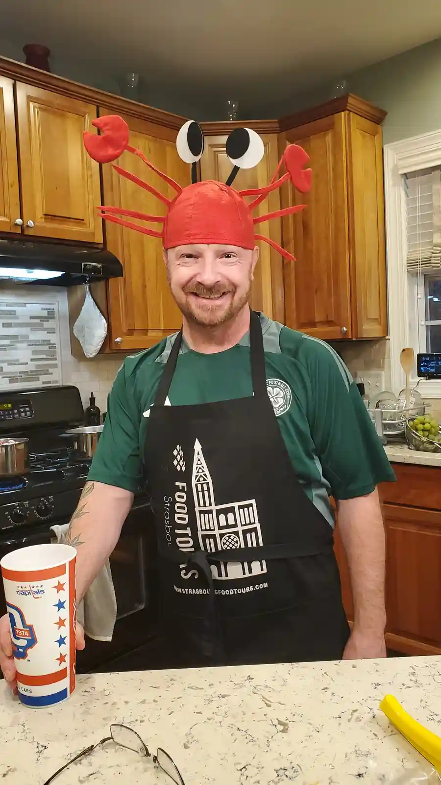 A smiling man in a kitchen wears a red lobster hat with googly eyes and claws, a black apron with a 'Strasbourg Food Tours'…