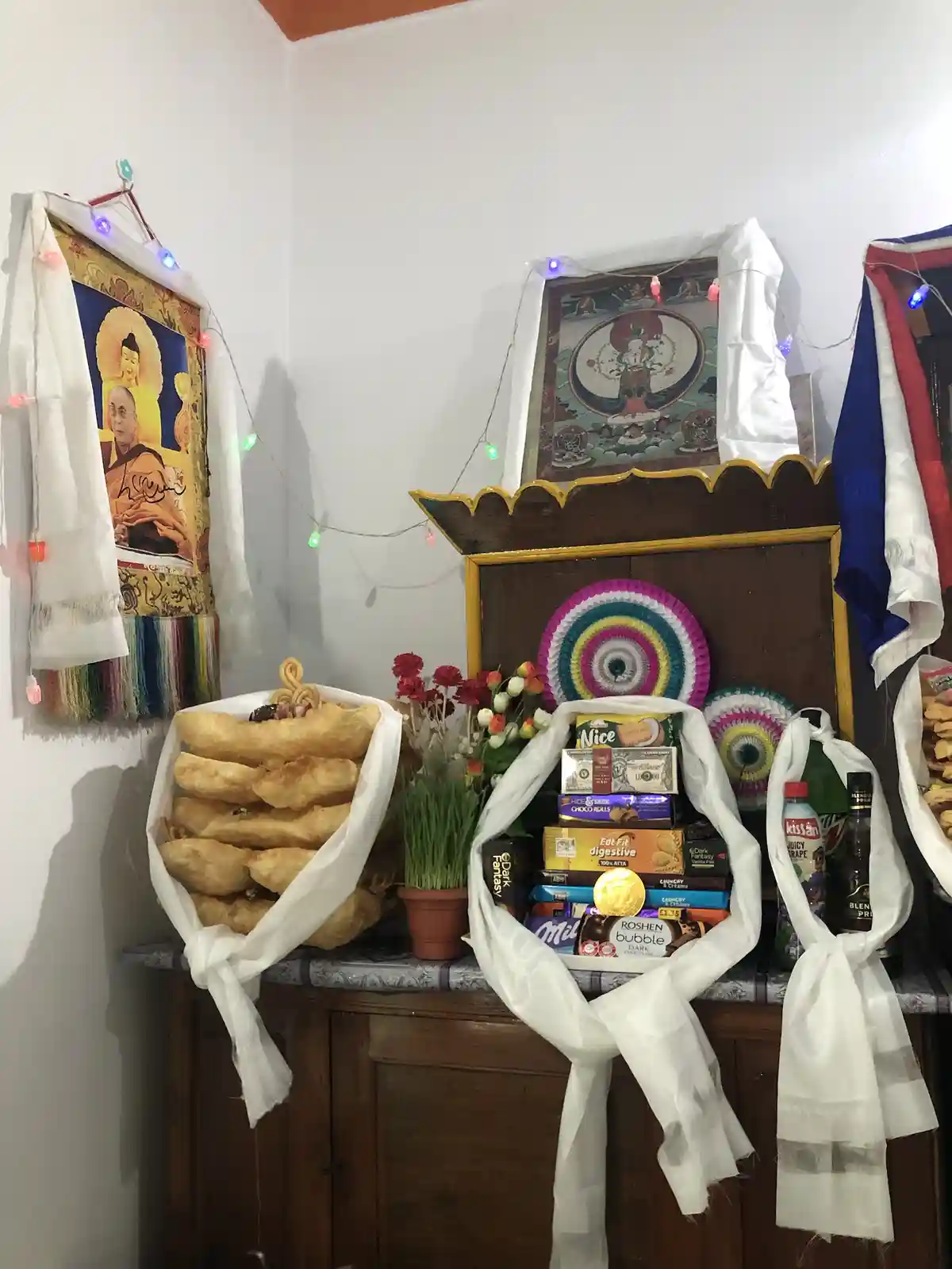 A wooden cabinet is adorned with religious Tibetan-style thangkas and colorful prayer flags, draped with white ceremonial…