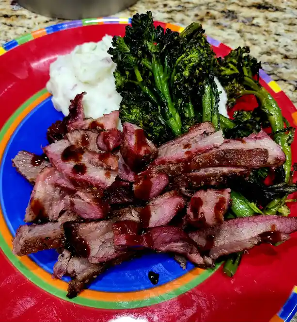 A colorful plate holds a meal of sliced medium-rare steak glazed with dark sauce, roasted broccoli with charred tips, and a…
