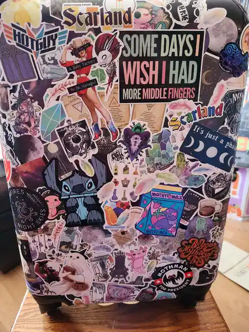 A black suitcase is covered in a dense collage of colorful stickers featuring fantasy, occult, and pop culture imagery like…
