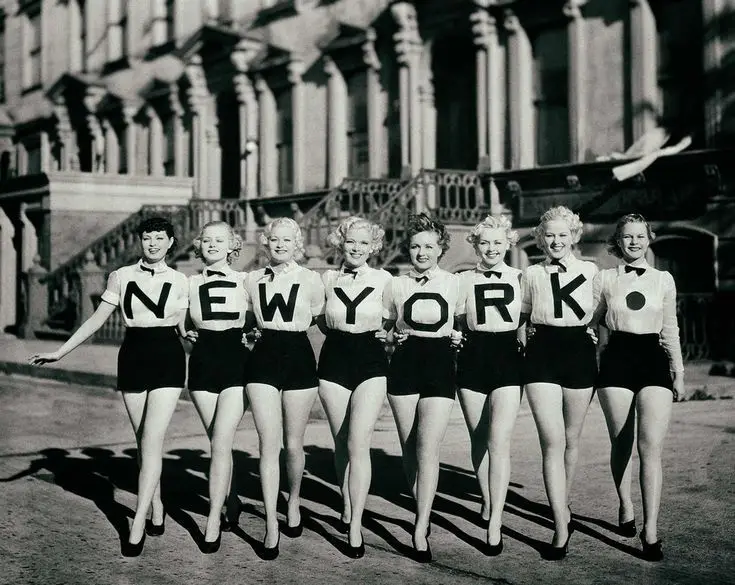 Seven vintage-style women in matching white shirts and black shorts stand in a line on a city street, each wearing a large…