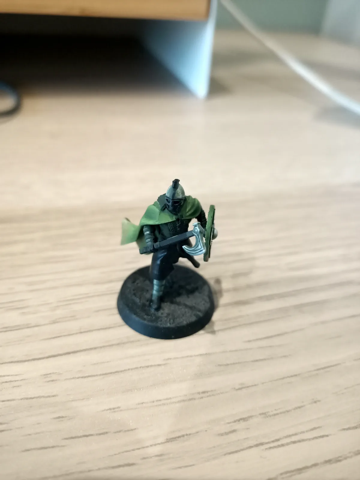 A painted miniature figurine of a warrior in green cloak and helmet, holding a shield and axe, stands on a black circular…
