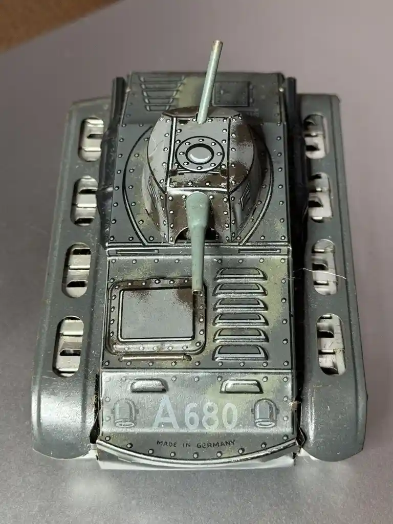 A top-down view of a vintage, silver-colored metal toy tank with a turret, cannon, and rivet details. The front of the tank…