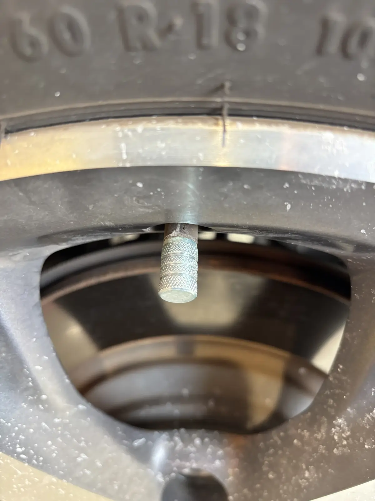A close-up photo shows a metallic, knurled valve stem protruding from the center of a tire's rim, with tire sidewall…