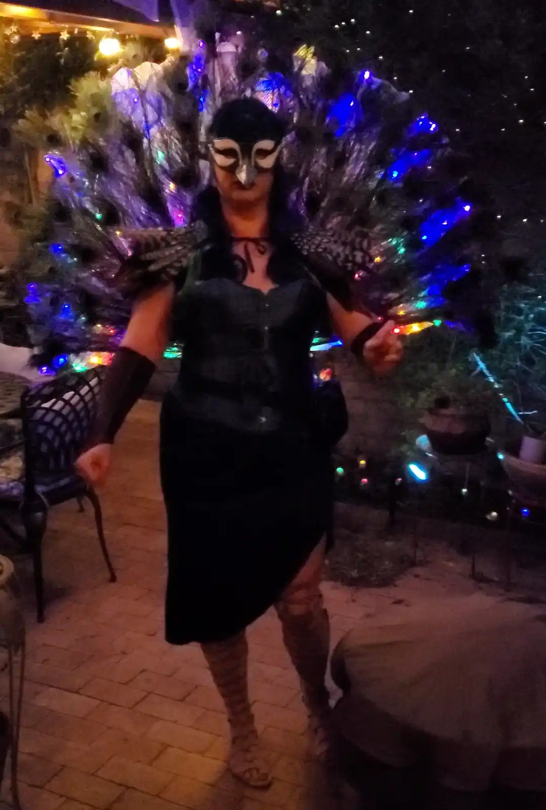 A person wearing a black peacock costume with a large, fanned-out tail made of feathers and colorful lights, along with a…