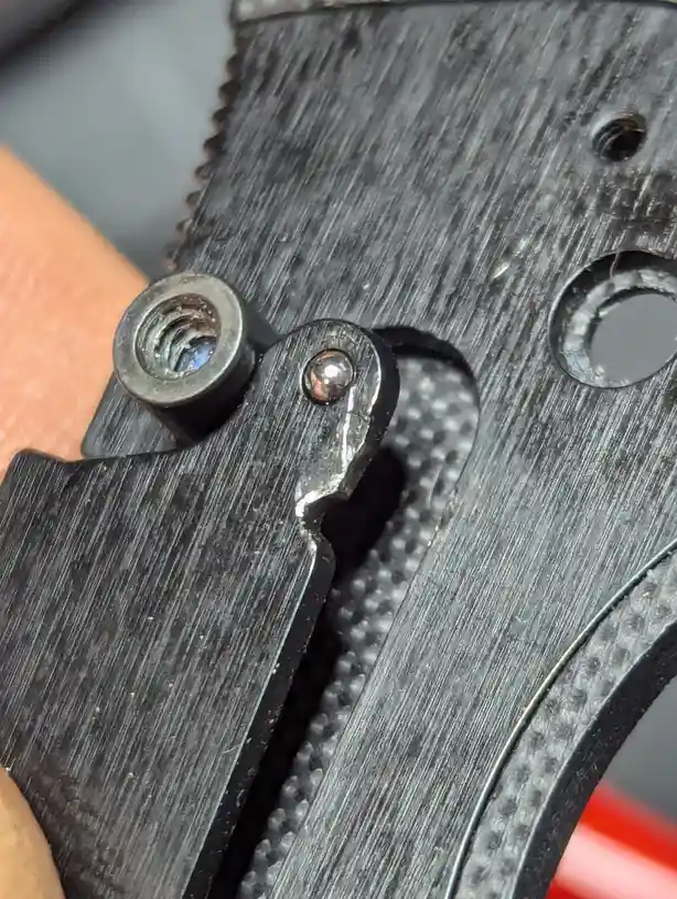 A close-up photo shows a black, textured folding knife's pivot mechanism, with a visible screw and a small, shiny ball…