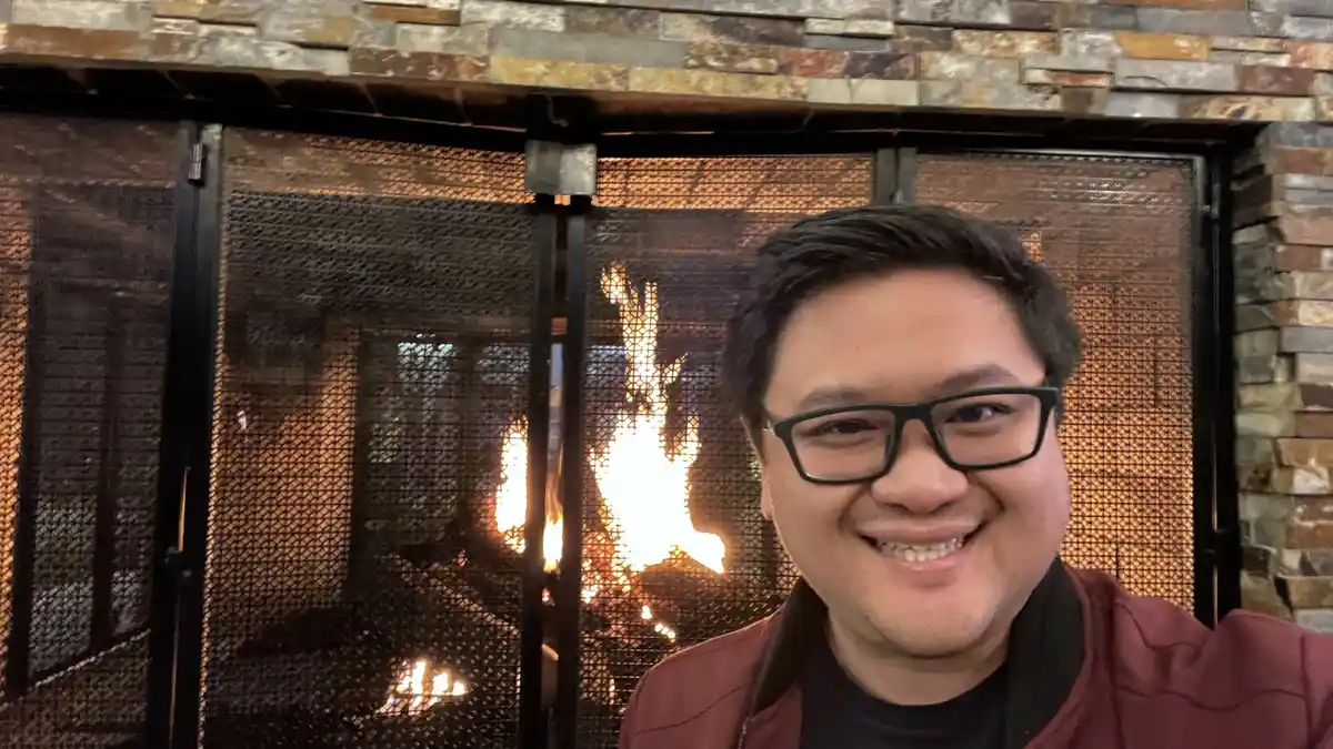A smiling man with glasses and a red jacket takes a selfie in front of a lit fireplace with a stone surround. The fire burns…