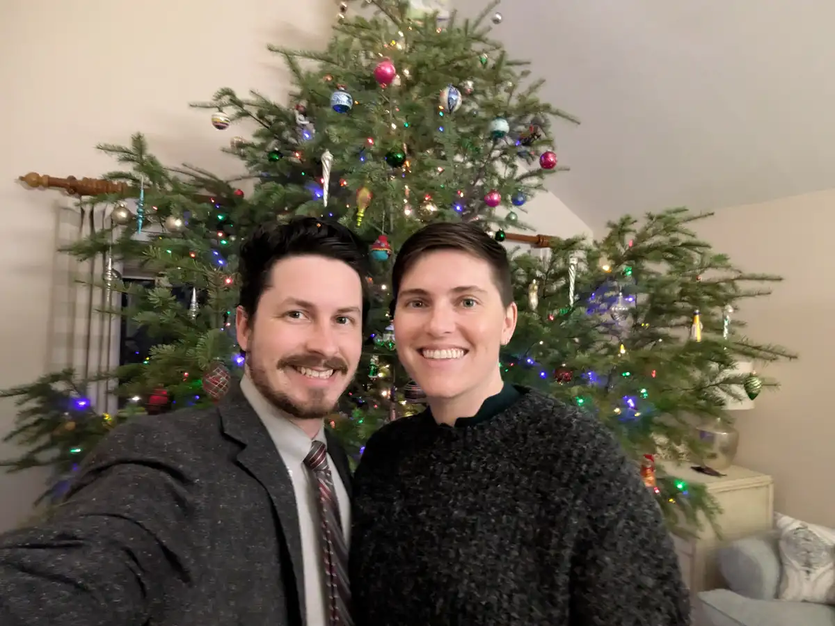 A selfie of a smiling man and woman standing in front of a decorated Christmas tree with colorful lights and ornaments. The…