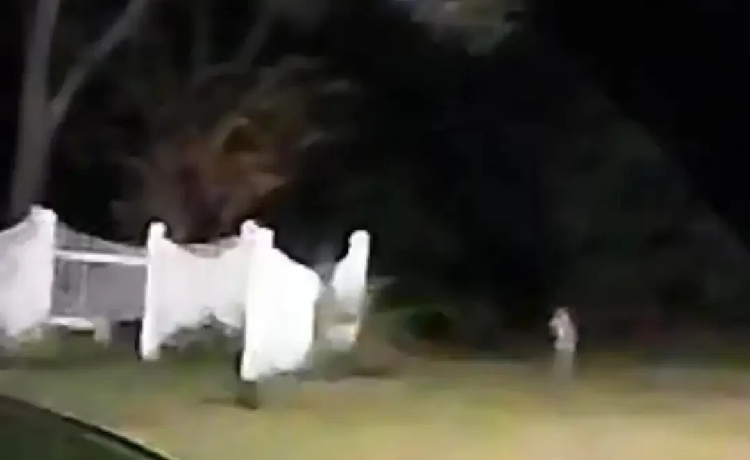A blurry, low-resolution night video shows a person standing in a yard near a white fence, with dark trees and foliage in…