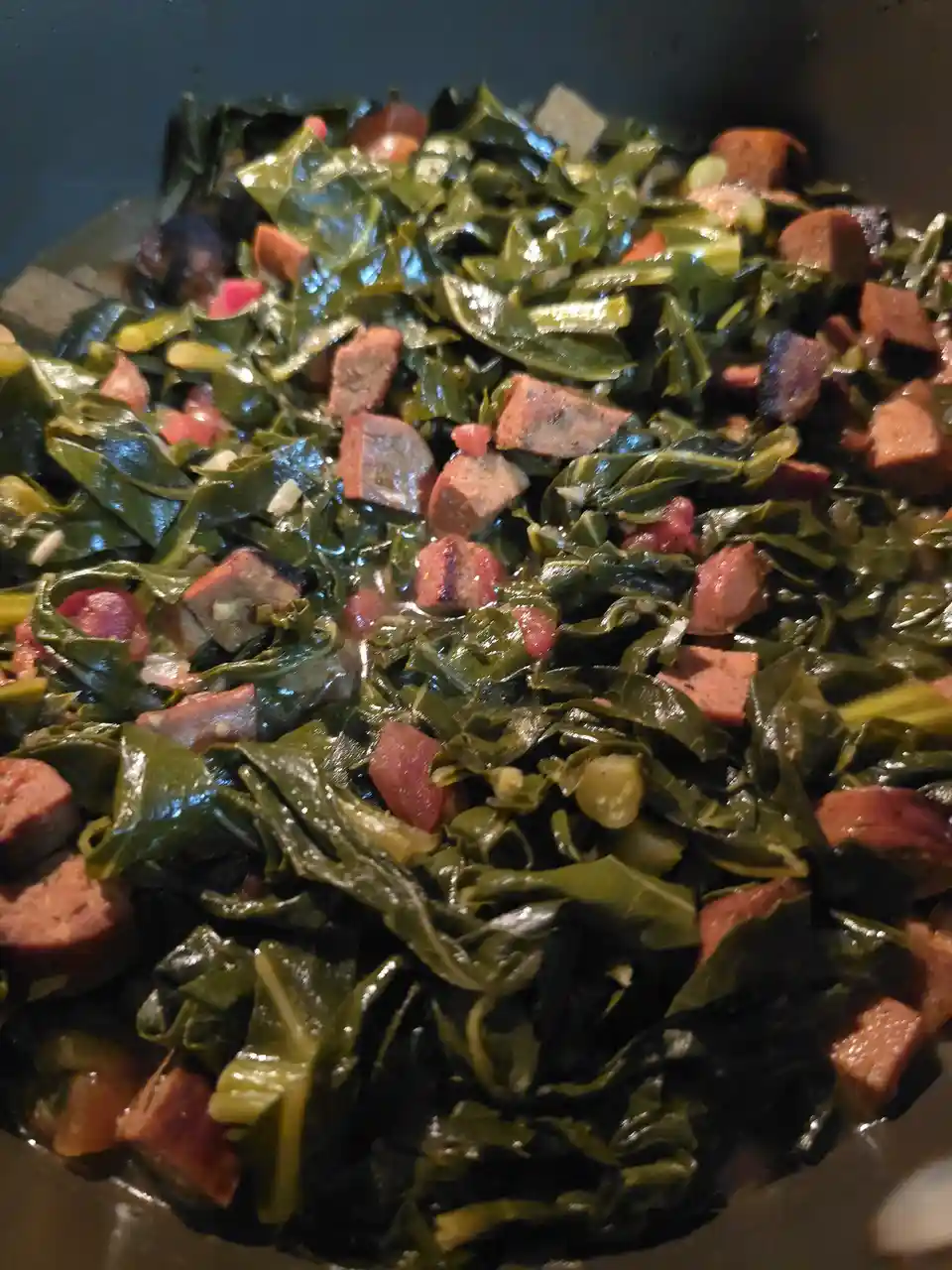 A close-up photo shows a pot filled with cooked collard greens mixed with chopped sausage pieces. The greens are dark green…