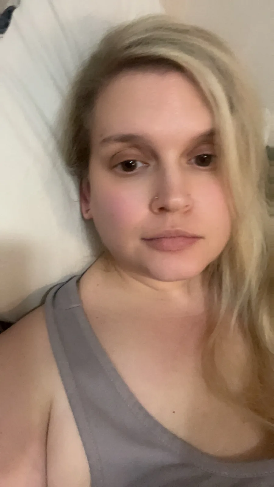 A person with blonde hair and a nose ring lies in bed wearing a gray tank top, looking directly at the camera with a neutral…