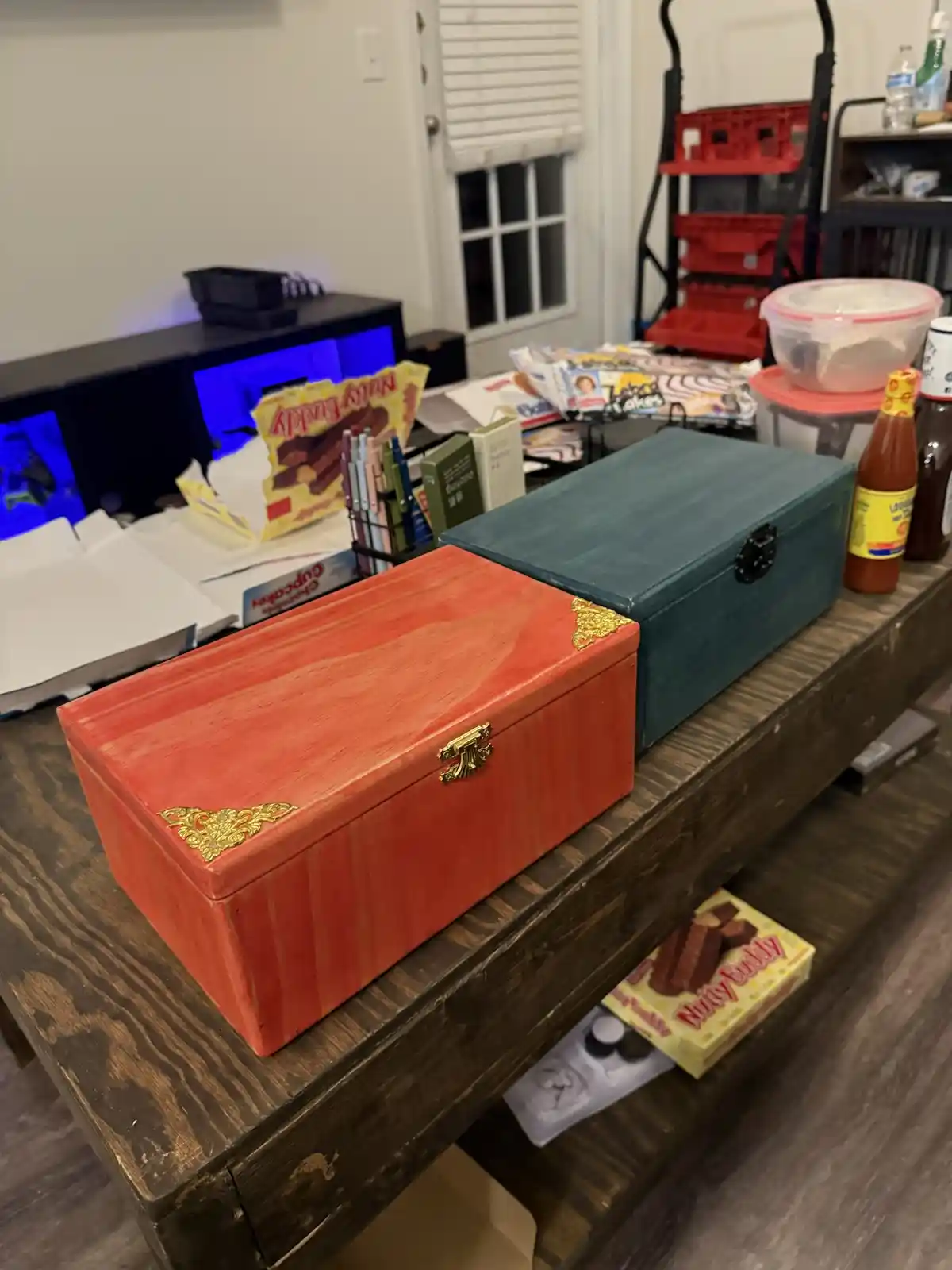 Two hand-painted wooden boxes, one red and one teal, sit side-by-side on a dark wooden table. The red box has ornate gold…