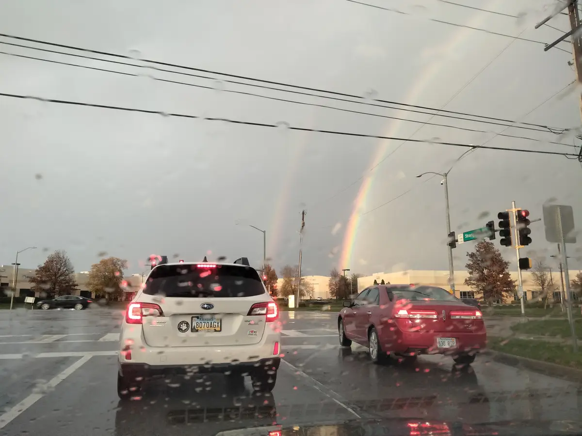 A photo taken from inside a car during rain, showing two cars stopped at a red light with a double rainbow visible in the…