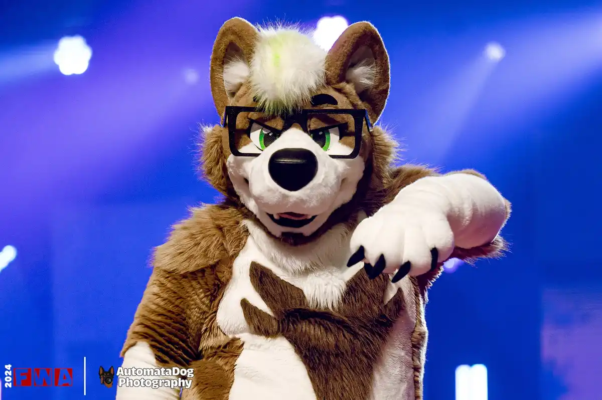 A person in a brown and white wolf fursuit with green eyes, black-rimmed glasses, and a green streak in their white forehead…