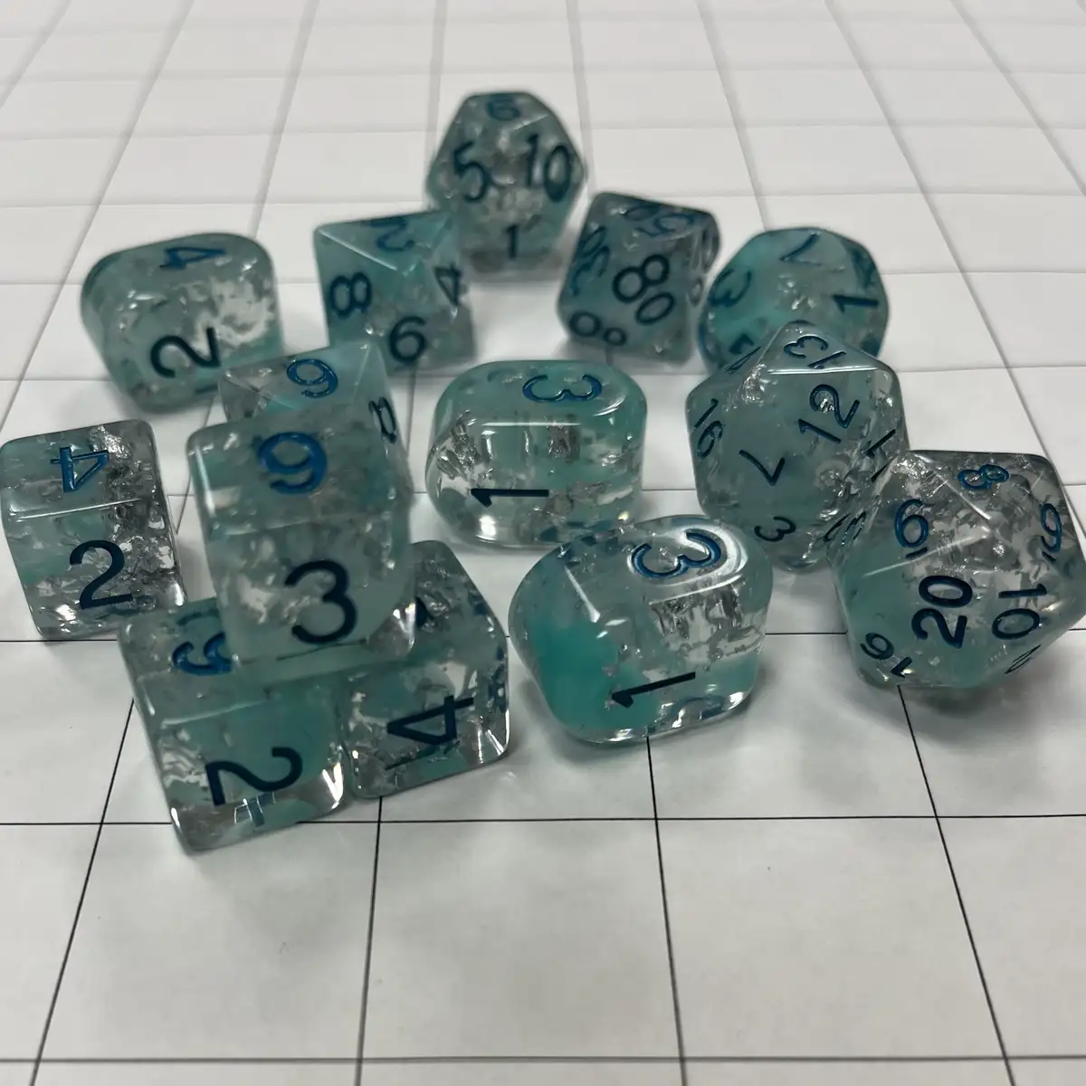 A collection of translucent teal and clear polyhedral dice with blue numbers are scattered on a white tiled surface, many…