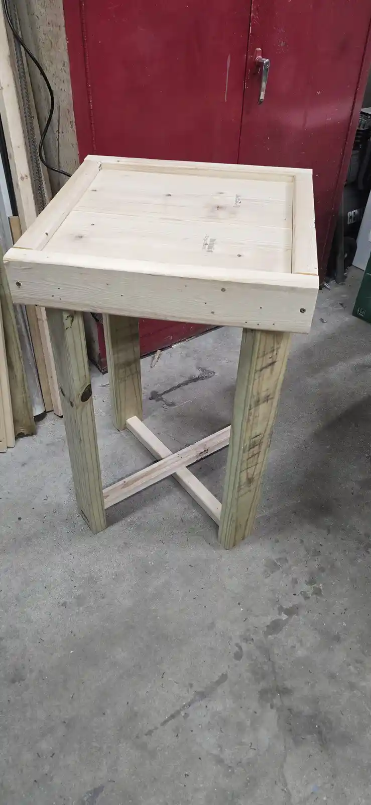 A crudely built wooden table with four legs and a cross-brace sits on a concrete floor in a workshop, with a red door in the…